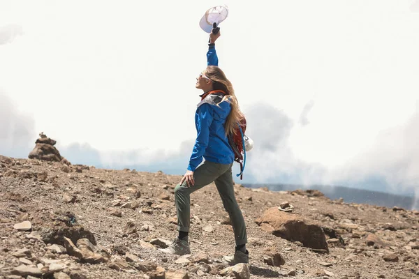 苗条勇敢的女孩在运动穿 蓝色夹克和靴子站在山上 地上的岩石 身后的云彩 举起她的手 握在拳头的帽子 背包与所有旅游物品 — 图库照片