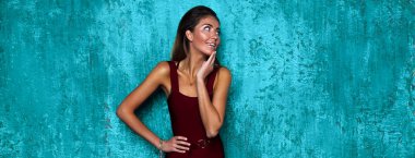 Bordo elbiseli genç bayanın modaya uygun portresi. O, bir el bel ve çene başka bir uzağa bakıyor. Şık makyaj ve hacim saç modeli. Boynundaki aksesuarlar. Mavi duvar arkasında