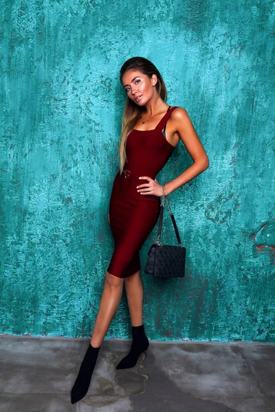 Stylish girl in red dress with open arms and waist belt. Hands on the waist, black purse hanging on wrist. High heel shoes, long legs. Pretty woman with modern makeup and volume hair.