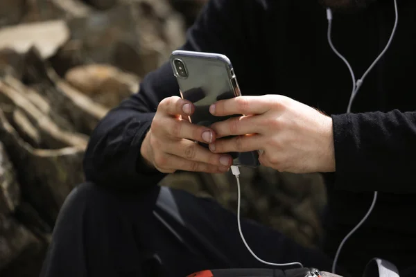 Close-up picture of black modern smartphone with white earphones. Man holding phone in hands and typing, searching in internet. Wearing black clothes.