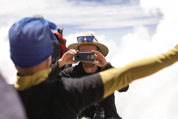 Two men having fun outdoor and posing for the cool pictures on the nature background. First guy holding his phone horizontal and taking pictures of his friend, clouds and mountains behind.