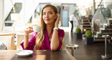 Bir kafede çekici genç model yaz sabahı çay içiyor. Güneş parlıyor, sıcak bir gün. Pembe ipek elbise giyiyor. Modern makyaj ve şık saç. Kulak dokunmak. Bilekten izle.