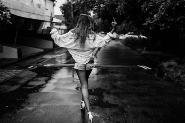 Cool young girl walking down the street on the high heels, wearing jeans shorts and oversize shirt. Tender skin, slim perfect body. Long pretty hair lying on the back. Black and white photo