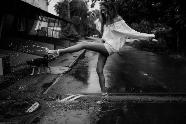 Attractive lady splashing water in a puddle with her tender leg in a high heel shoes. Funny and crazy woman. Stylish romantic look. Wet after rain hair and clothes. Slim body with tender skin