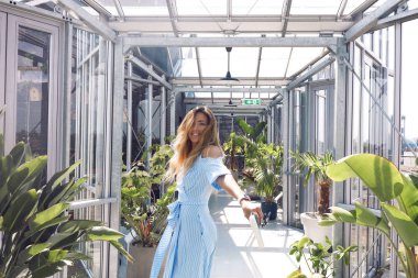 Mutlu model gülümsüyor ve yeşil evde tek başına eğleniyor. Bitkilerin ve çiçeklerin yanında dans ediyor ve yürüyor. İnce vücudunda süslü, uzun bir elbise. Aksesuarlar ve yüzünde şık bir makyaj, dalgalı saçlar.