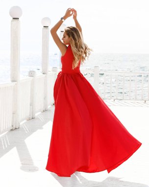 Mükemmel görünümlü hanımefendinin üzerinde yumuşak, kırmızı bir elbise. Güneşin altında dışarıda duruyor ve denize bakıyor. Ellerini havaya kaldırdı, rüzgar elbisesini salladı ve uzun sarı saçları vardı..   