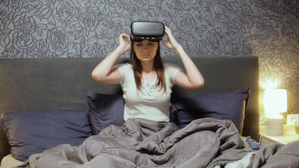 Красивая молодая женщина в VR гарнитуре на кровати. Оглядываясь вокруг, улыбаясь и используя жесты руками. Смотреть видео, играть в игру . — стоковое видео