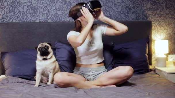 Bella giovane donna che indossa VR Headset seduta sul letto con cane carlino. Guardati intorno. Guarda il video VR, gioca a VR . — Video Stock