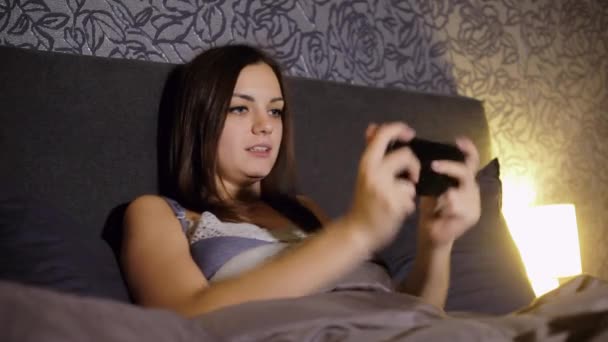 Mujer joven jugando juegos en el teléfono inteligente mientras está acostado en la cama en casa por la noche. Tensely juega, gana el juego — Vídeo de stock