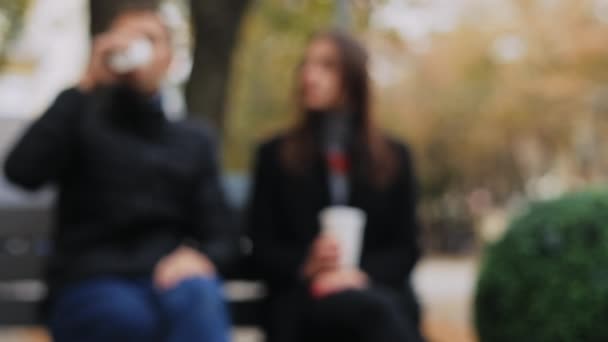 Sfondo sfocato, coppia felice con bicchieri di carta in mano che parla sulla panchina della città per strada — Video Stock