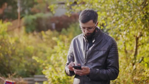 Молодий бородатий чоловік використовує телефон у сонячному парку — стокове відео