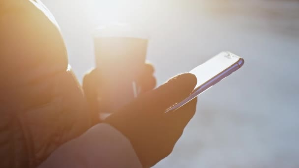 Close up of the girls hands in gloves, using smartphone and drinking coffee, sunny winter, — Stock Video