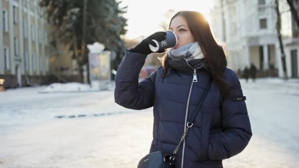 Gyönyörű fiatal nő ital forró kávét egy papír poharat a napsütéses téli, havas város utca — Stock videók