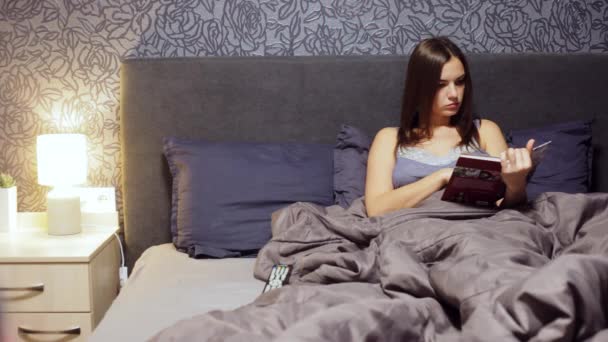 Hermosa joven acostada en la cama en el dormitorio, leer un libro culinario. Mujer descansando en la noche en casa . — Vídeos de Stock