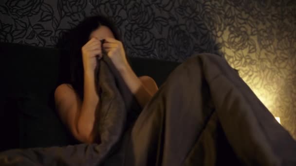 Uma jovem assiste a um filme de terror na TV enquanto está deitada na cama, com medo e cobre seu rosto — Vídeo de Stock