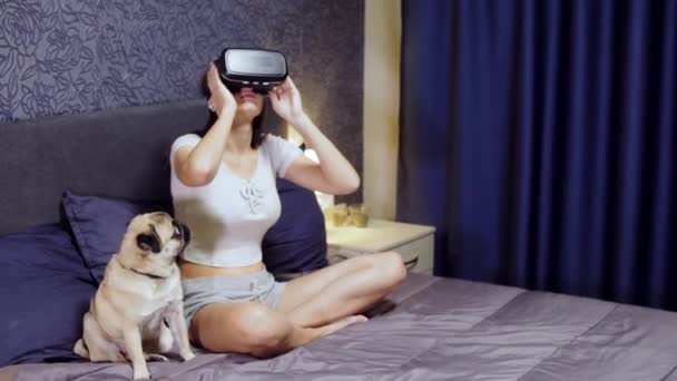 Hermosa mujer joven con VR Headset sentado en la cama con perro de pug. Mirando alrededor. Ver vídeo VR, juego VR juego . — Vídeo de stock
