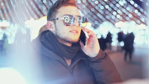 Hombre elegante hablando por teléfono en la noche de Navidad cerca del árbol de Año Nuevo de la ciudad principal, un reflejo de las luces en gafas — Vídeos de Stock