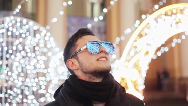 Stylish bearded man at celebrate the new year, city Christmas tree, garlands and lights reflected in the mirrored sunglasses — Stock Video