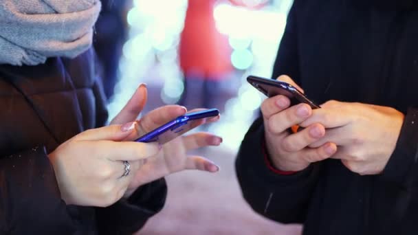 Zbliżenie dłoni mężczyzny i kobiety używa aplikacji mobilnej w śnieżny wieczór, chating z przyjaciółmi, w pobliżu światła i dekoracji na Boże Narodzenie — Wideo stockowe