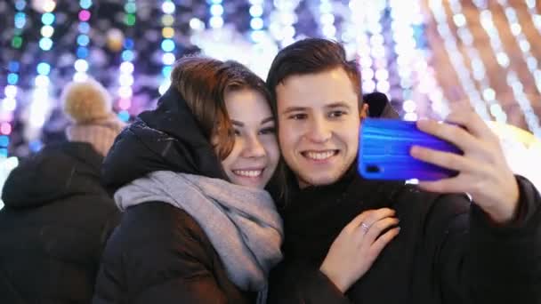 Счастливая пара делает селфи на современном телефоне, улыбаясь и целуясь в новогоднюю ночь — стоковое видео