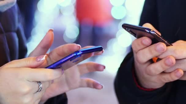 Primer plano de la mano del hombre y la mujer utiliza el teléfono móvil en la noche nevada, chating con amigos, luces y decoraciones para la Navidad en el bacground . — Vídeos de Stock