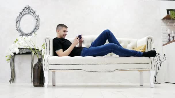 若い男はソファでリラックスとモバイル ゲームをプレイ モダンで豪華な家でスマート フォンを使用して — ストック動画