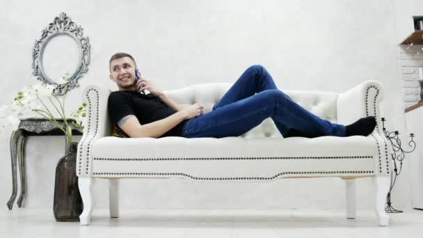 Młody człowiek, relaksując się na kanapie i mówi przez telefon komórkowy w nowoczesny, luksusowy dom — Wideo stockowe