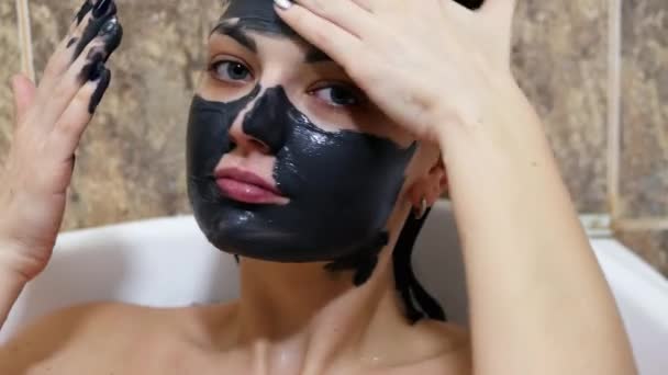 Mulher coloca no rosto máscara creme de limpeza preta e olhando para a câmera. Conceito de cuidados da pele — Vídeo de Stock