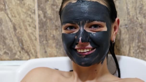 Frau mit schwarzer Reinigungscreme-Maske im Gesicht blickt in die Kamera und lächelt. Hautpflegekonzept — Stockvideo