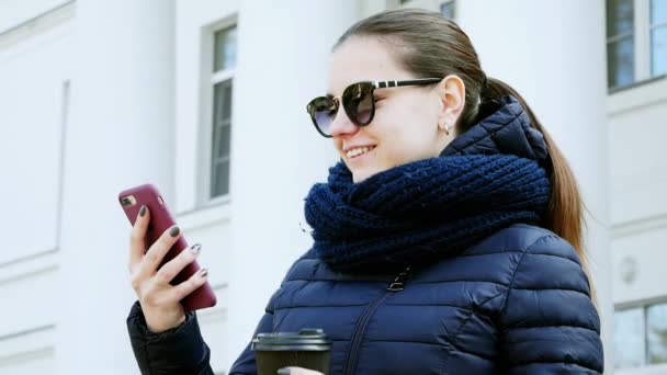Κοντινό-up πορτραίτο μιας νεαρής γυναίκας στα ποτήρια πίνοντας καφέ χρησιμοποιώντας το έξυπνο τηλέφωνο στην πόλη — Αρχείο Βίντεο