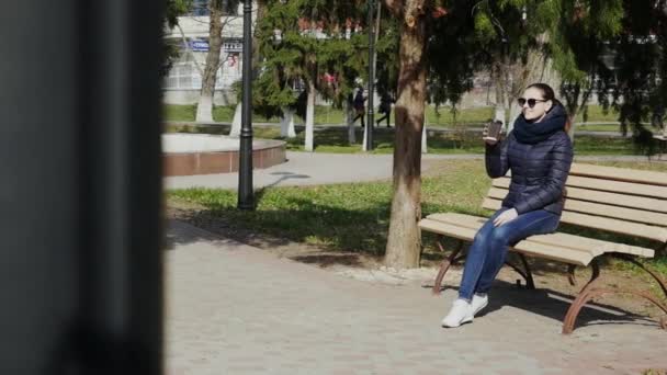 Молодая женщина пьет кофе на скамейке в солнечном парке — стоковое видео