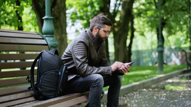 Bearded man använder telefonen i parken på bänken — Stockvideo