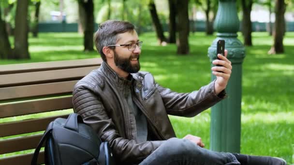 ハンサムなあごひげの男の笑顔、電話でのビデオ通話、公園で. — ストック動画