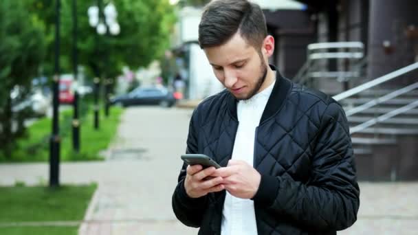 Bonito jovem barbudo usa o telefone na rua, digitando texto — Vídeo de Stock