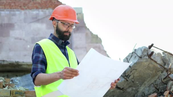 Бородатый инженер-строитель в оранжевом шлеме на разрушенном фоне здания. Серьезный строитель анализирует рисунок. Подготовка к строительству — стоковое видео