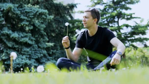 Man blowing paardebloem zittend in een stadspark op het gras, rust in de stad — Stockvideo