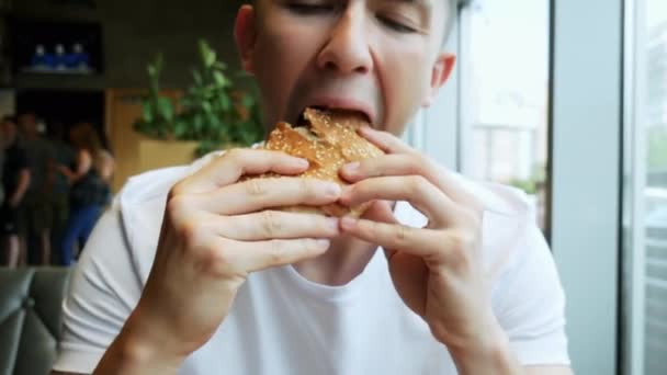 Nahaufnahme von jungen hungrigen Mann isst Hamburger in einem Café, Fast-Food-Restaurant — Stockvideo