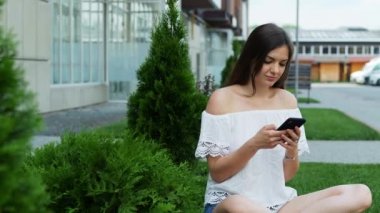 Genç ve güzel bir kadın otelin yakınındaki çimenlikte otururken telefonu kullanıyor.