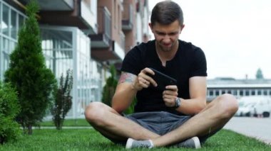Mutlu adam akıllı telefon mobil oyun oynamak, evin yakınındaki çim üzerinde oturan.