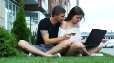 Mutlu çift dizüstü bilgisayar kullanarak online alışveriş yapar, mal seçin ve binanın yakınındaki çim üzerinde oturan bir kredi kartı numarası girer