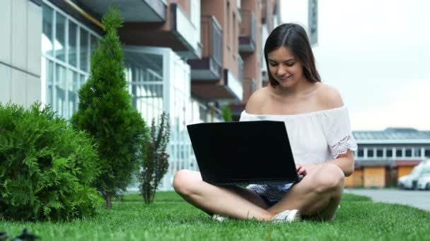 Молодая красивая деловая женщина, печатающая на ноутбуке, сидящая на траве возле отеля, женщина пользуется ноутбуком в парке — стоковое видео