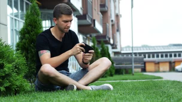 Ung man spelar mobilspel på smartphone, sitter på gräset nära byggnaden. — Stockvideo