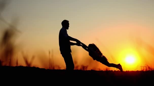 Силуэты отца и дочери, держащиеся за руки и вращающиеся на прекрасном закате в поле — стоковое видео
