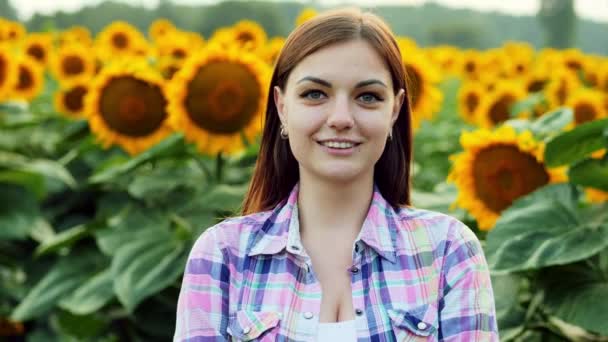 Hermosa joven agricultora está mirando a la cámara y sonriendo en el fondo de un campo con girasoles — Vídeo de stock