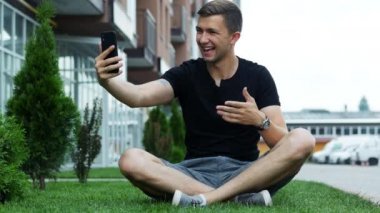 Yakışıklı genç adam cep telefonu ile görüntülü sohbet konuşma, çim üzerinde oturan ve arkadaşları ile konuşmak