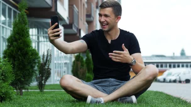 Stilig ung man pratar i videochatt via mobiltelefon, sitter på gräset och pratar med vänner — Stockvideo