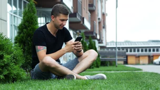 Uomo attraente utilizzare il telefono cellulare mentre seduto sull'erba vicino all'hotel, chiacchierando con gli amici — Video Stock