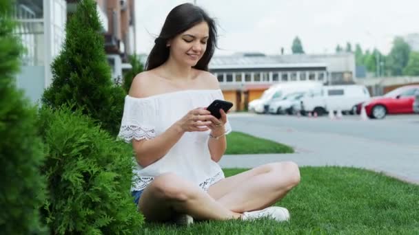 Jonge mooie vrouw gebruik slimme telefoon terwijl zittend op het gras in de buurt van het Hotel — Stockvideo