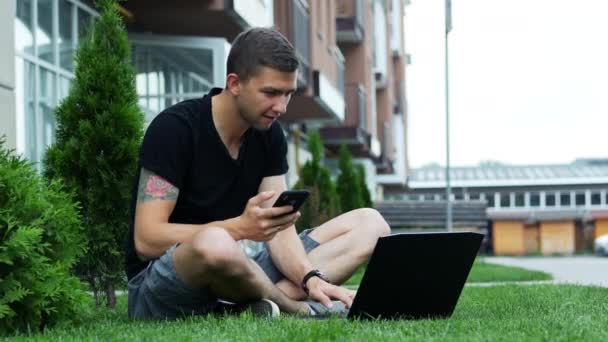 Mężczyzna uczeń używa telefonu i laptopa siedząc na trawie — Wideo stockowe