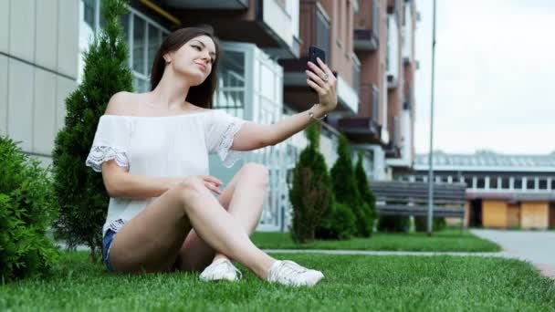 Schöne junge stilvolle Frau macht Selfie im Park, sitzt auf dem Gras — Stockvideo